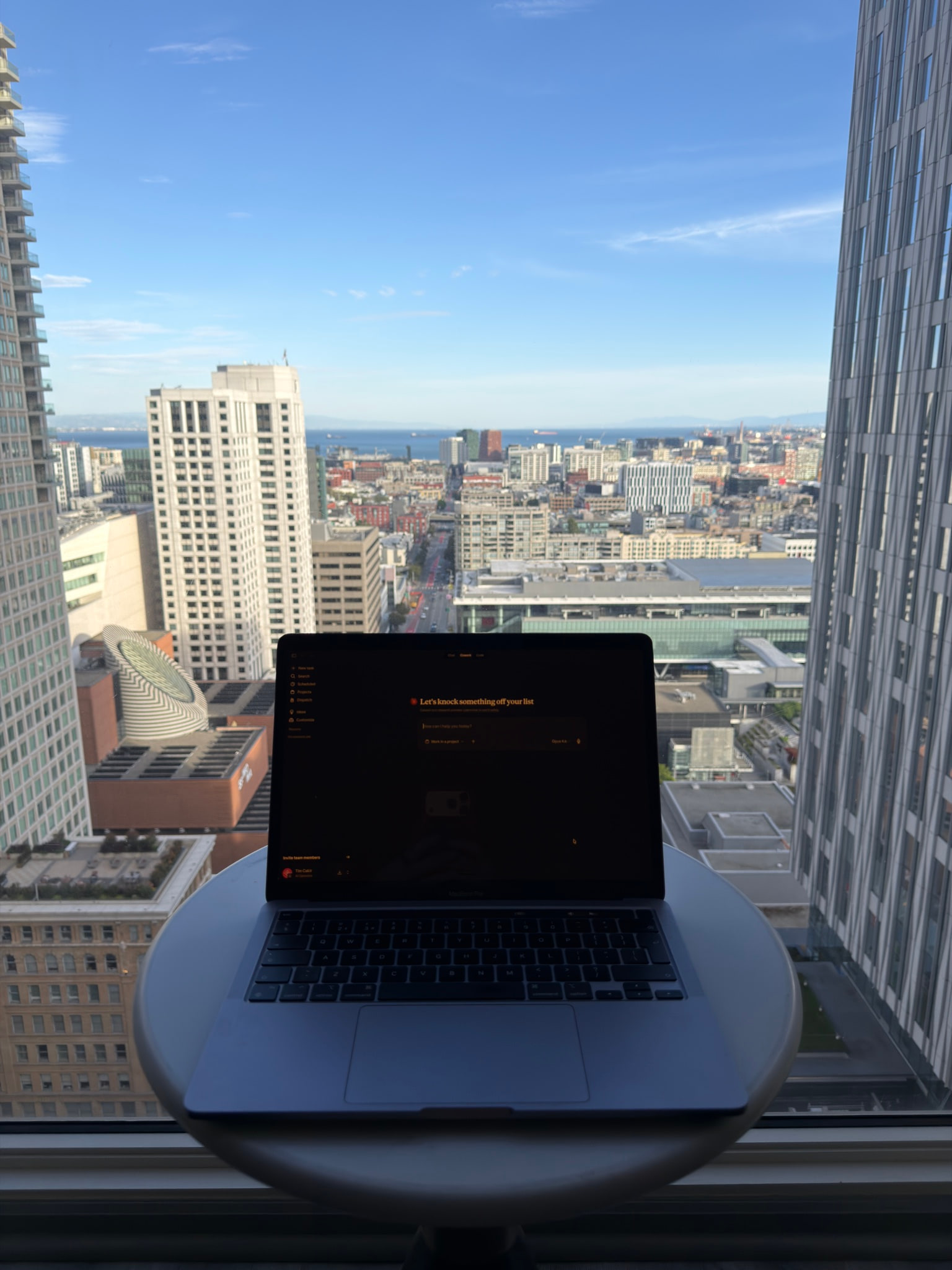 A laptop open on Claude, standing on a small round table in front of a floor-to-ceiling window, looking out over the San Francisco skyline towards the bay.