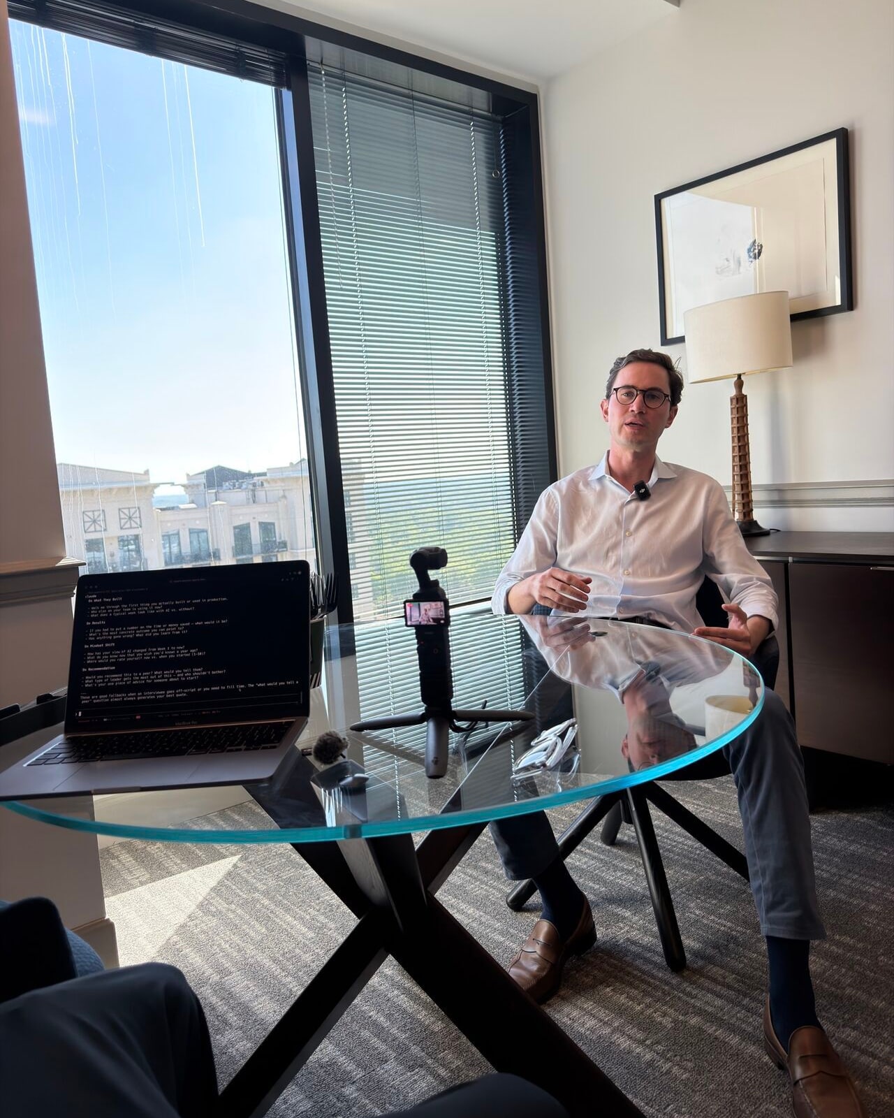 A customer seated at a glass table talking to camera during a filmed interview, with a tripod-mounted camera and a laptop of interview questions in front of him.