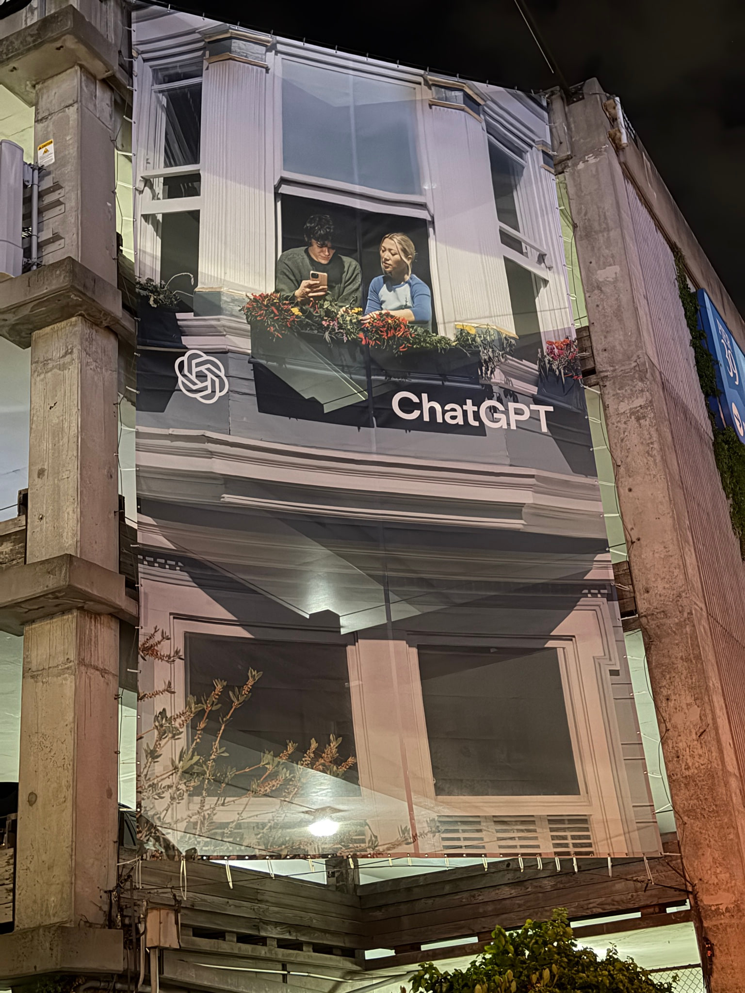 A building in San Francisco at dusk wrapped in a large ChatGPT advertisement showing two people leaning out of a window over a flower box.