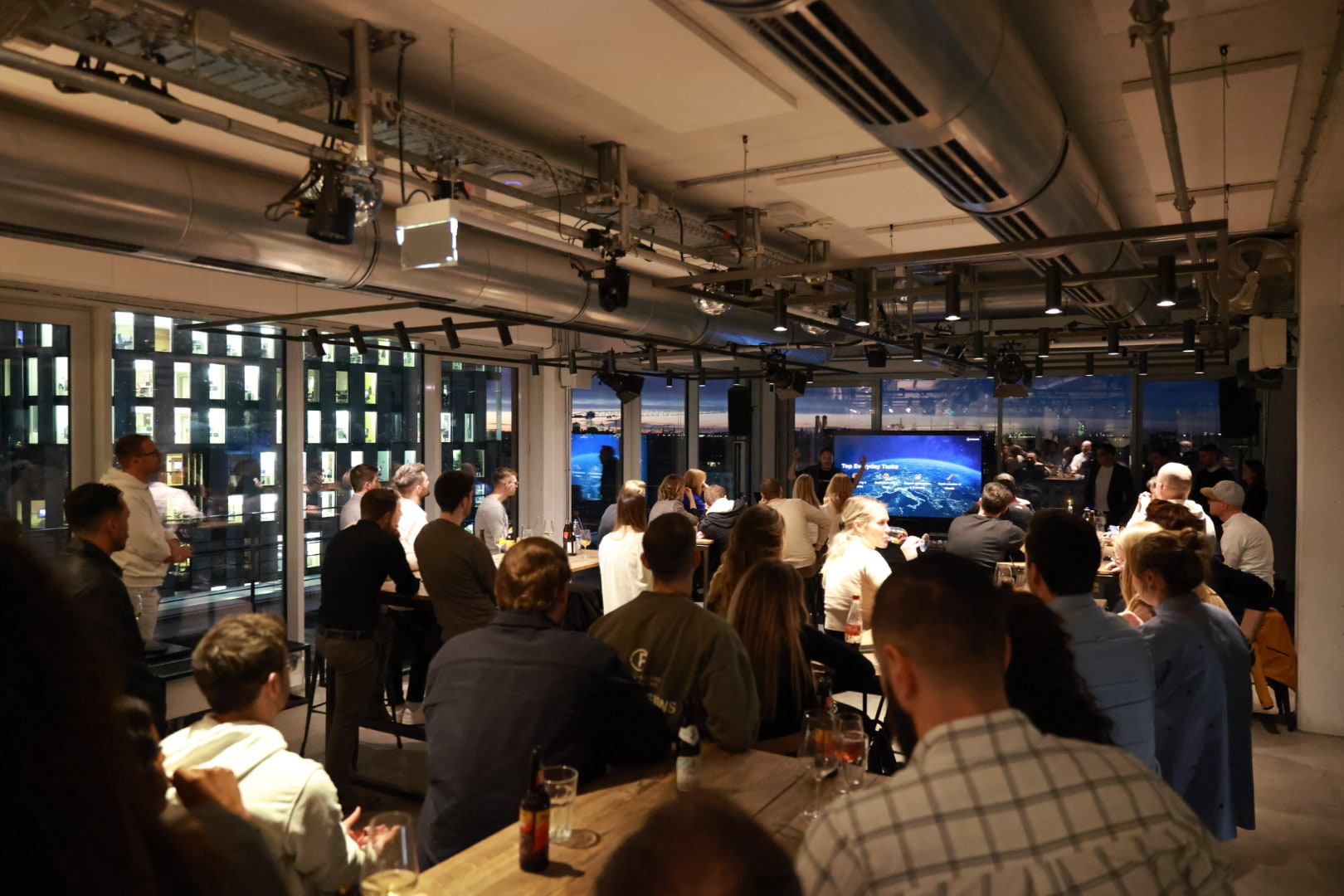 A full room of client team members at long tables during an evening session at WERK1 in Munich, a large screen lit at the far end.