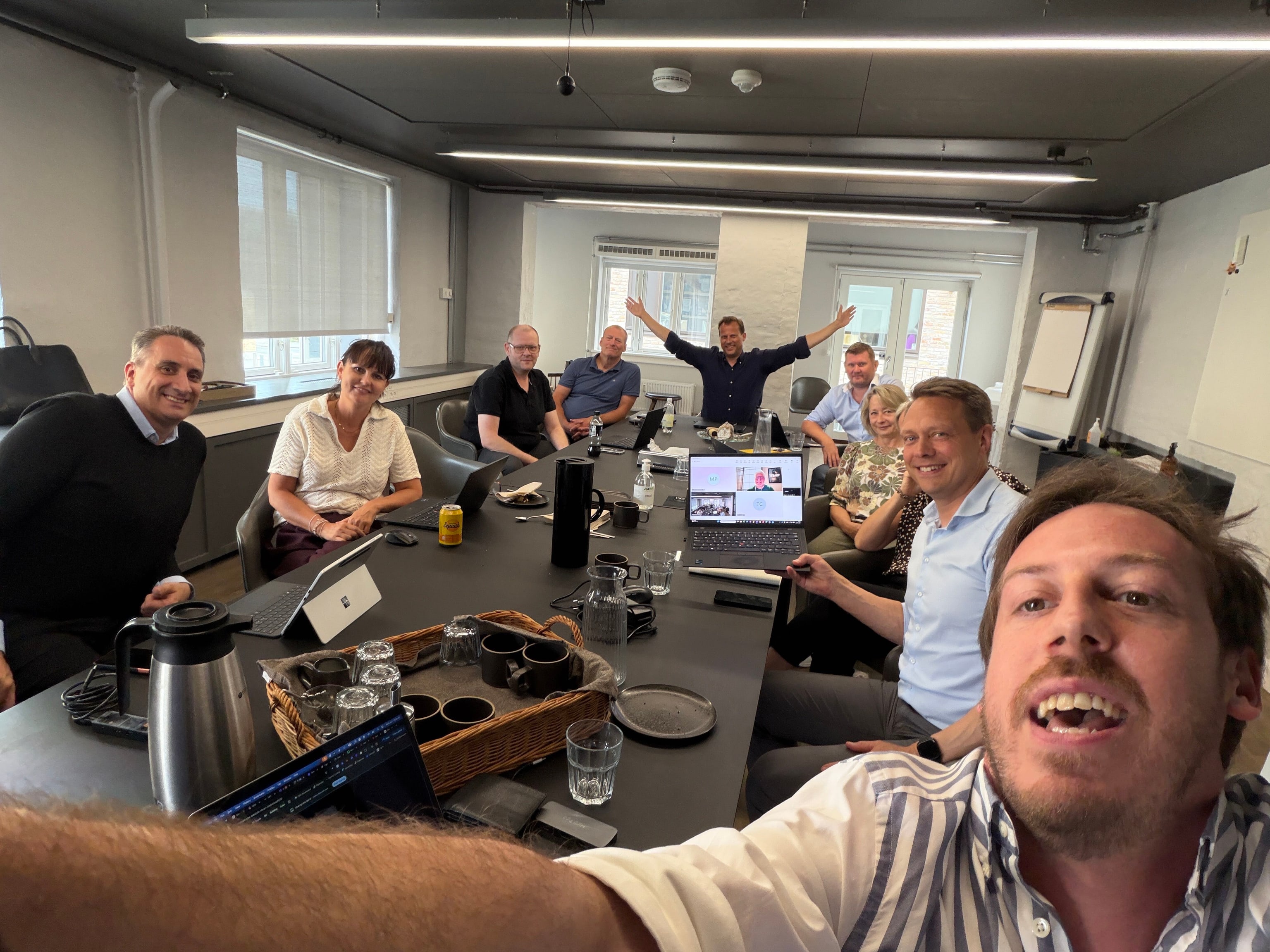 A client team around a boardroom table on a hybrid AI Operator session, one man standing with his arms thrown wide, Tim Cakir laughing in the foreground, and two more participants joining on a laptop screen in the middle of the table.