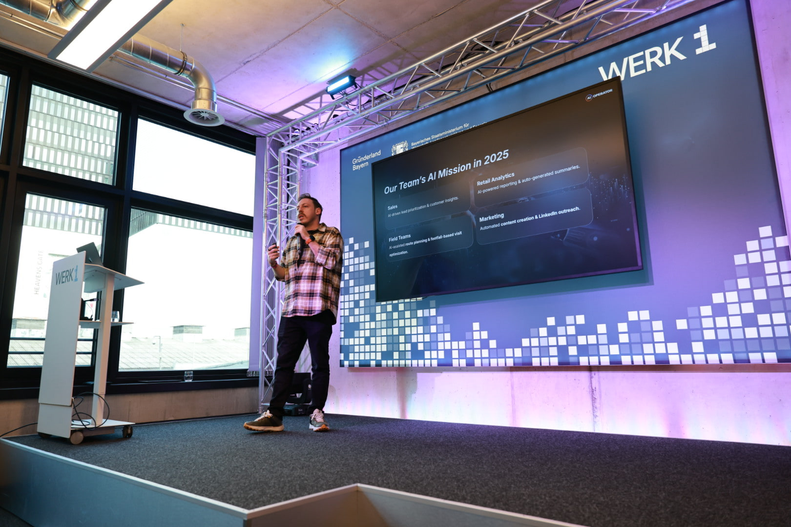 Tim Cakir on stage at WERK1 in Munich beside a large screen headed Our Team's AI Mission in 2025, addressing a client team.