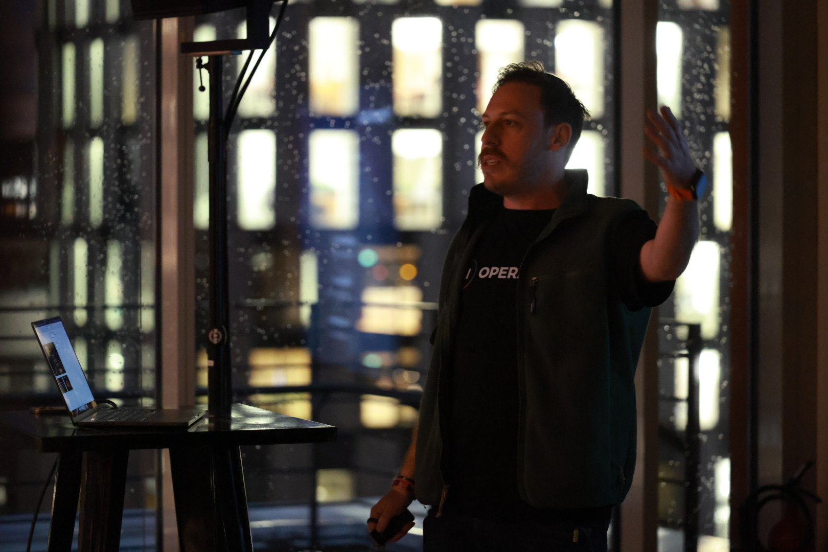 Tim Cakir mid-sentence with one hand raised, presenting from beside a laptop on a side table, with rain on the floor-to-ceiling windows and city lights behind him.