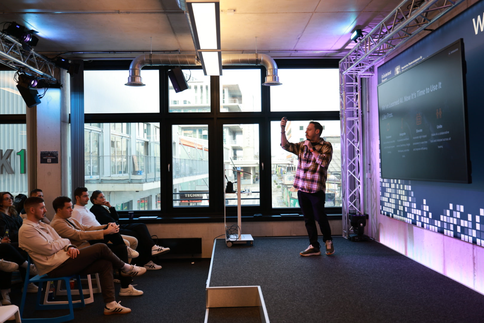 Tim Cakir on stage at WERK1 in Munich, one arm raised, addressing a seated client team with a lit screen beside him.